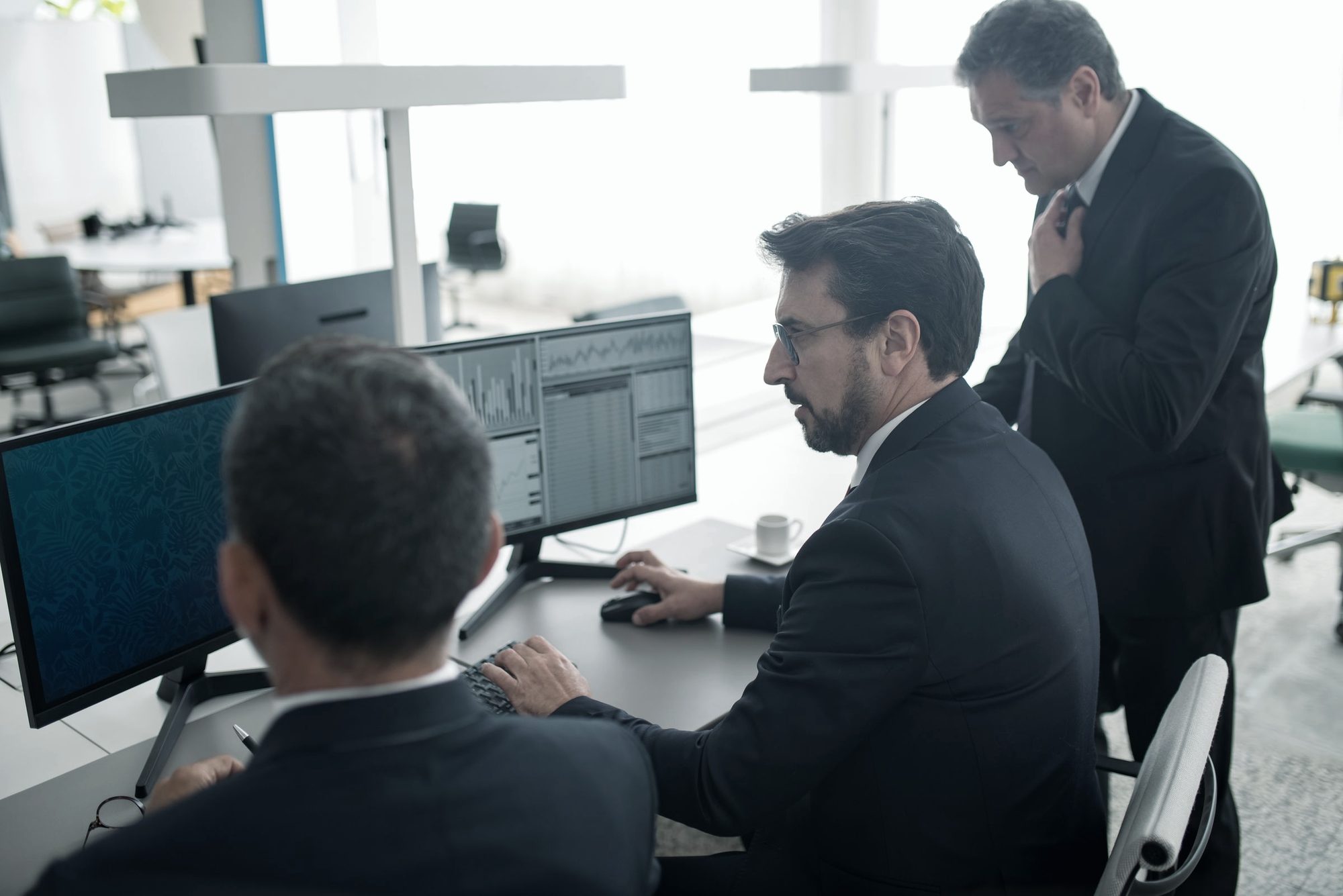 Two colleagues in suits reviewing work at a trading-floor desk while a senior colleague looks on
