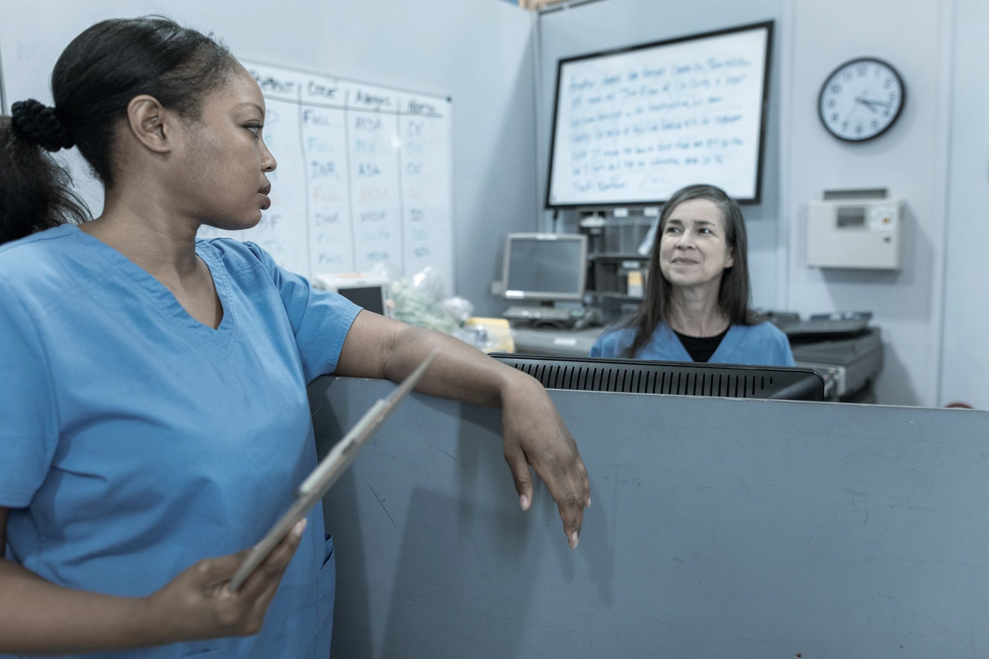 A nurse with a clipboard at a hospital nursing station, a colleague at the desk behind
